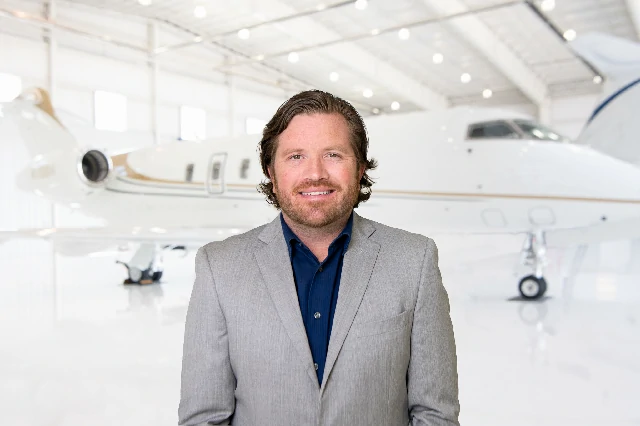 Smiling man with wavy brown hair wearing a light gray suit jacket and dark blue shirt, standing in front of a private jet inside a bright airplane hangar.