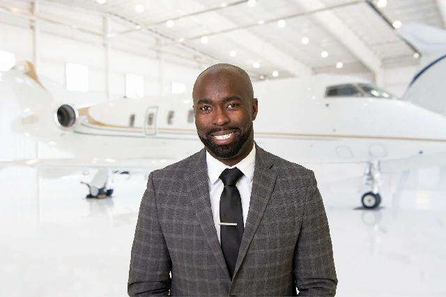 Smiling man with a beard wearing a gray checkered suit, white shirt, and black tie, standing in front of a private jet inside a bright airplane hangar.