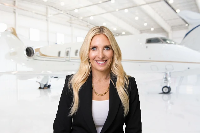 Smiling woman with long blonde hair wearing a black blazer and white top, standing in front of a private jet inside a bright airplane hangar.