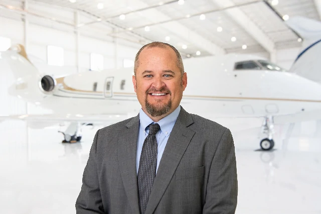 Smiling man in a gray suit and tie standing in front of a private jet inside a bright airplane hangar.
