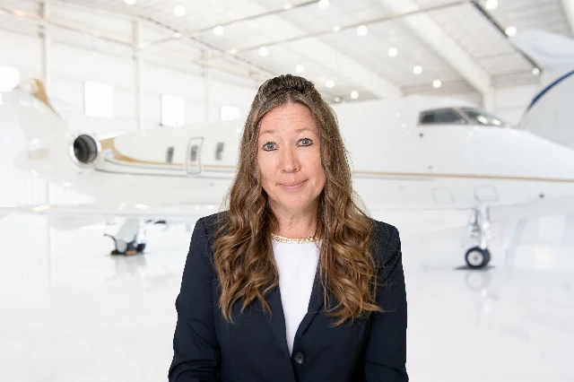 Smiling woman in a dark blazer and white blouse standing in front of a private jet inside a bright airplane hangar.