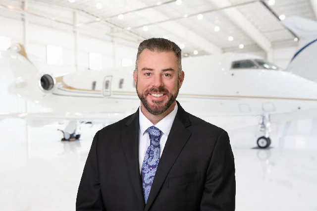 Smiling man in a dark suit and patterned tie standing in front of a private jet inside a bright airplane hangar.