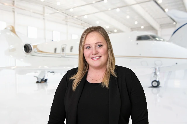 Smiling woman with straight blonde hair wearing a black blazer over a black top, standing in front of a private jet inside a brightly lit airplane hangar.