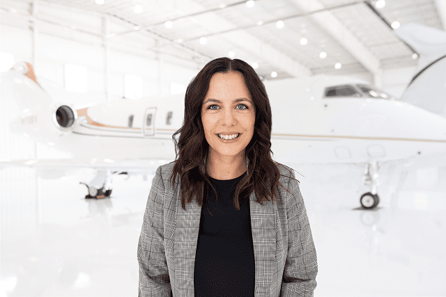 Woman with shoulder-length dark hair wearing a gray plaid blazer and black top, smiling while standing in a bright aircraft hangar with a white private jet in the background.